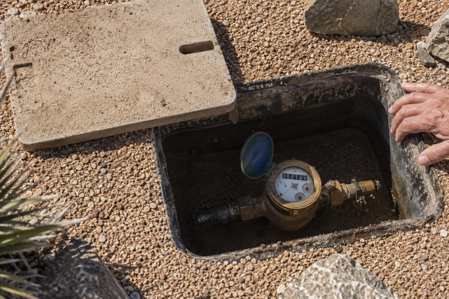 Open water meter box in a gravel yard with hands checking the meter dial for movement